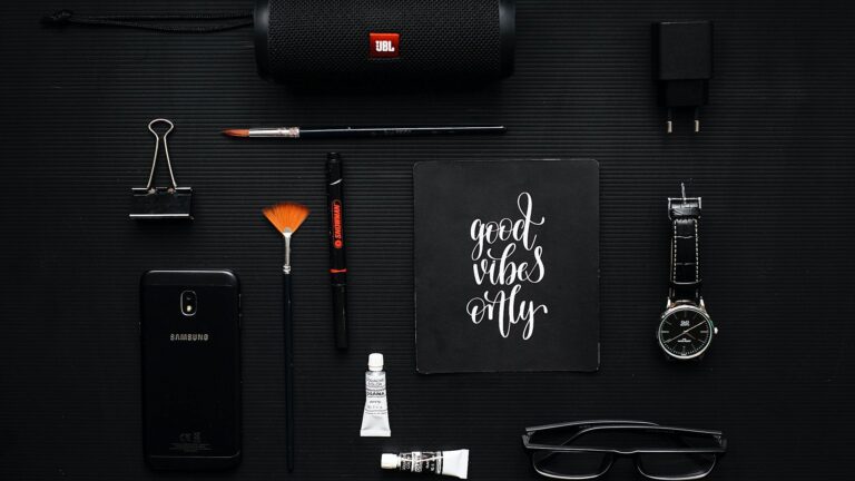 close up photography of smartphone beside binder clip speaker and eyeglasses on black textile